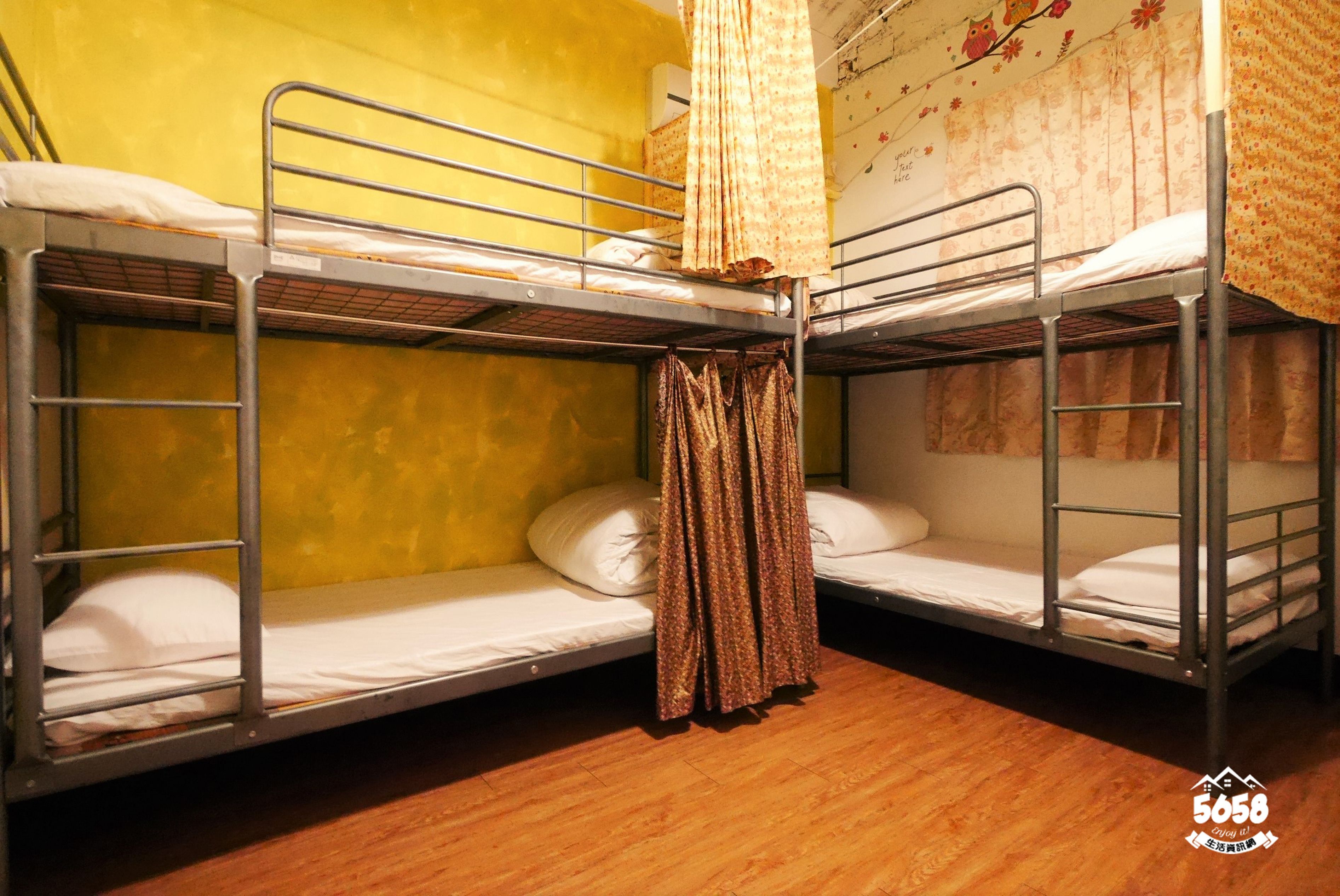 A dormitory room with bunk beds, wooden flooring, and a curtain on one bed.
