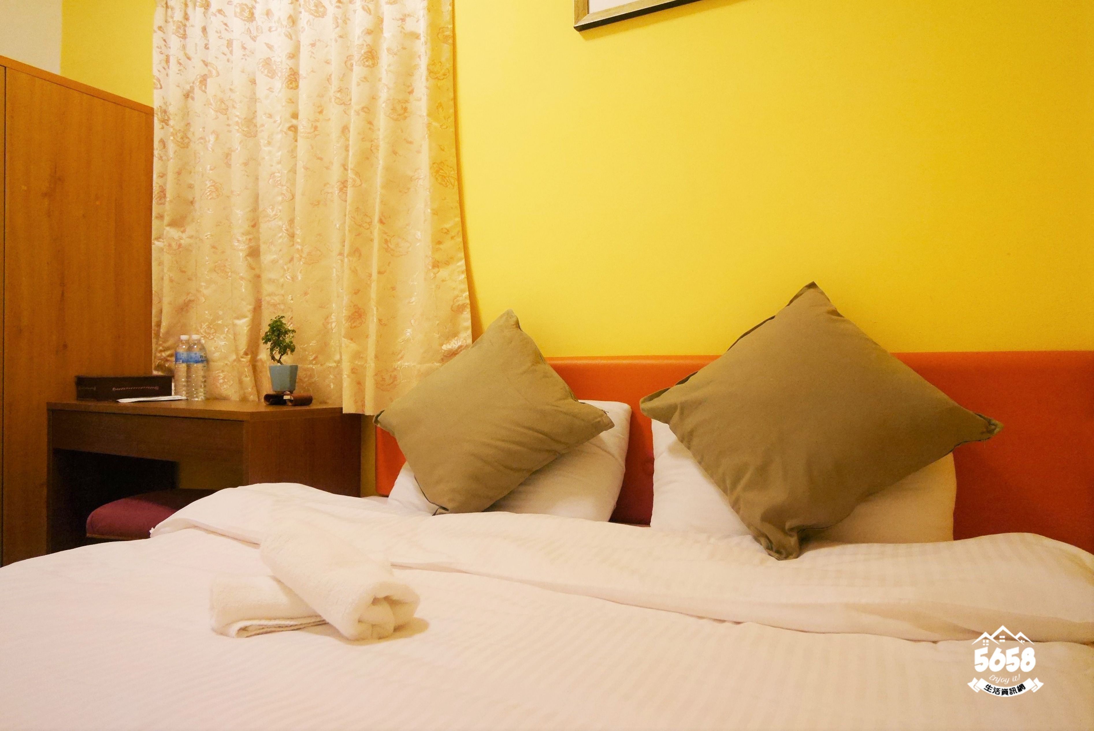 A hotel room with a bed, pillows, a nightstand, a plant, and a curtain.