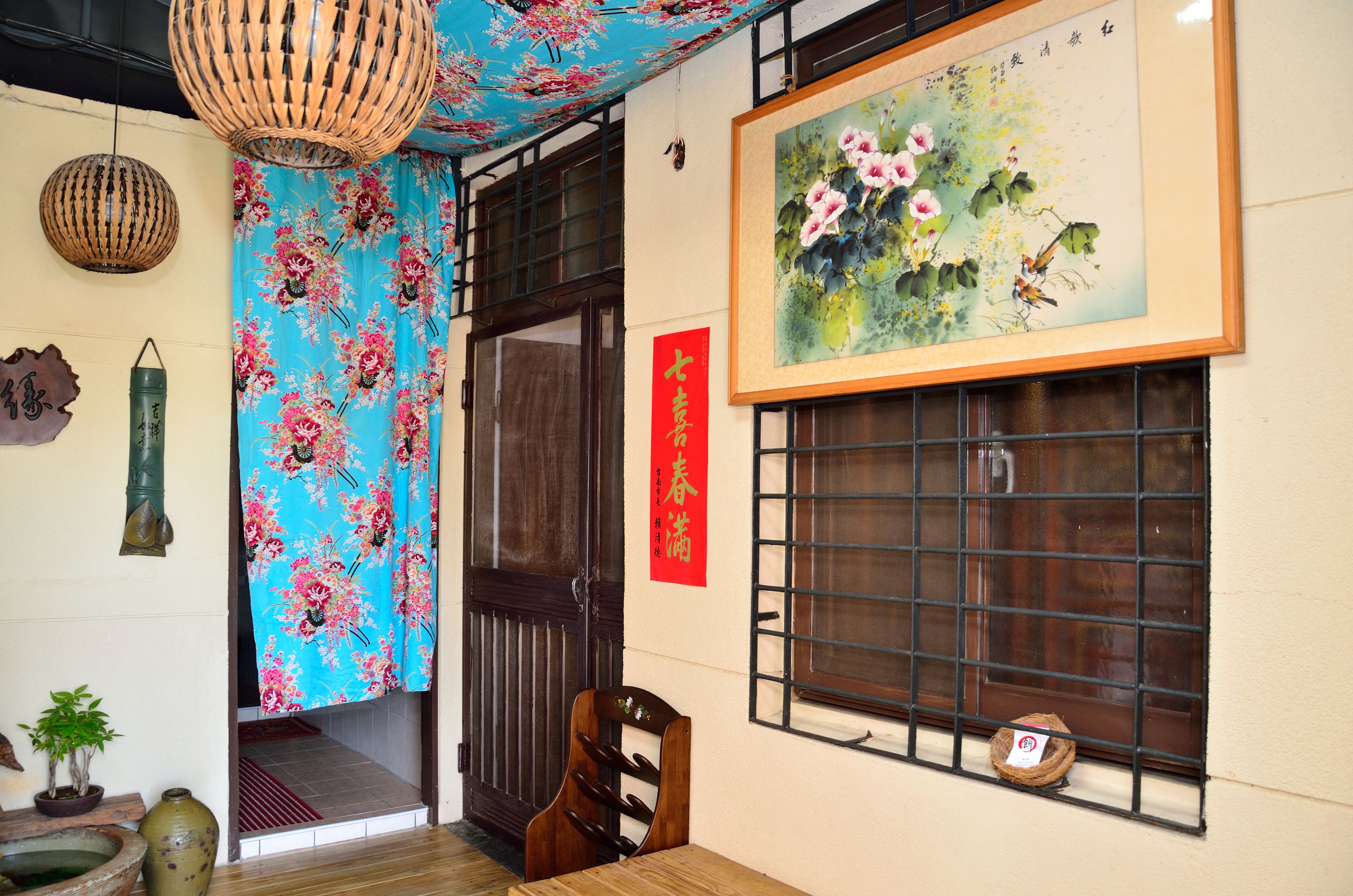 A traditional room with a wooden door, a window with metal bars, a hanging lantern, a framed painting, and a red vertical sign.