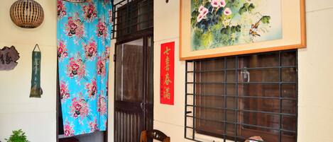 A traditional room with a wooden door, a window with metal bars, a hanging lantern, a framed painting, and a red vertical sign.