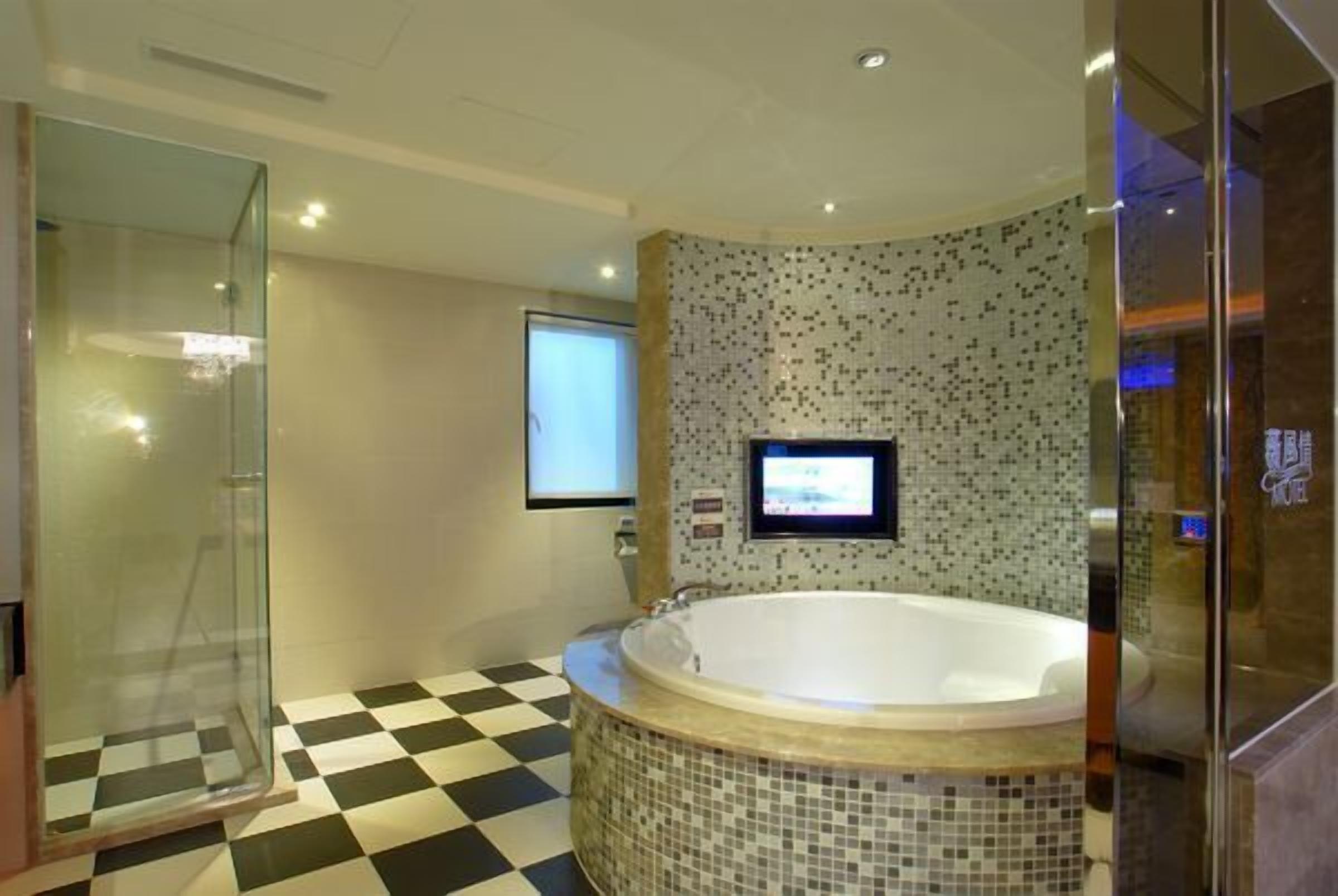 A modern bathroom with a checkered floor, a glass shower enclosure, a large freestanding bathtub, and a tiled wall with a screen mounted above it.