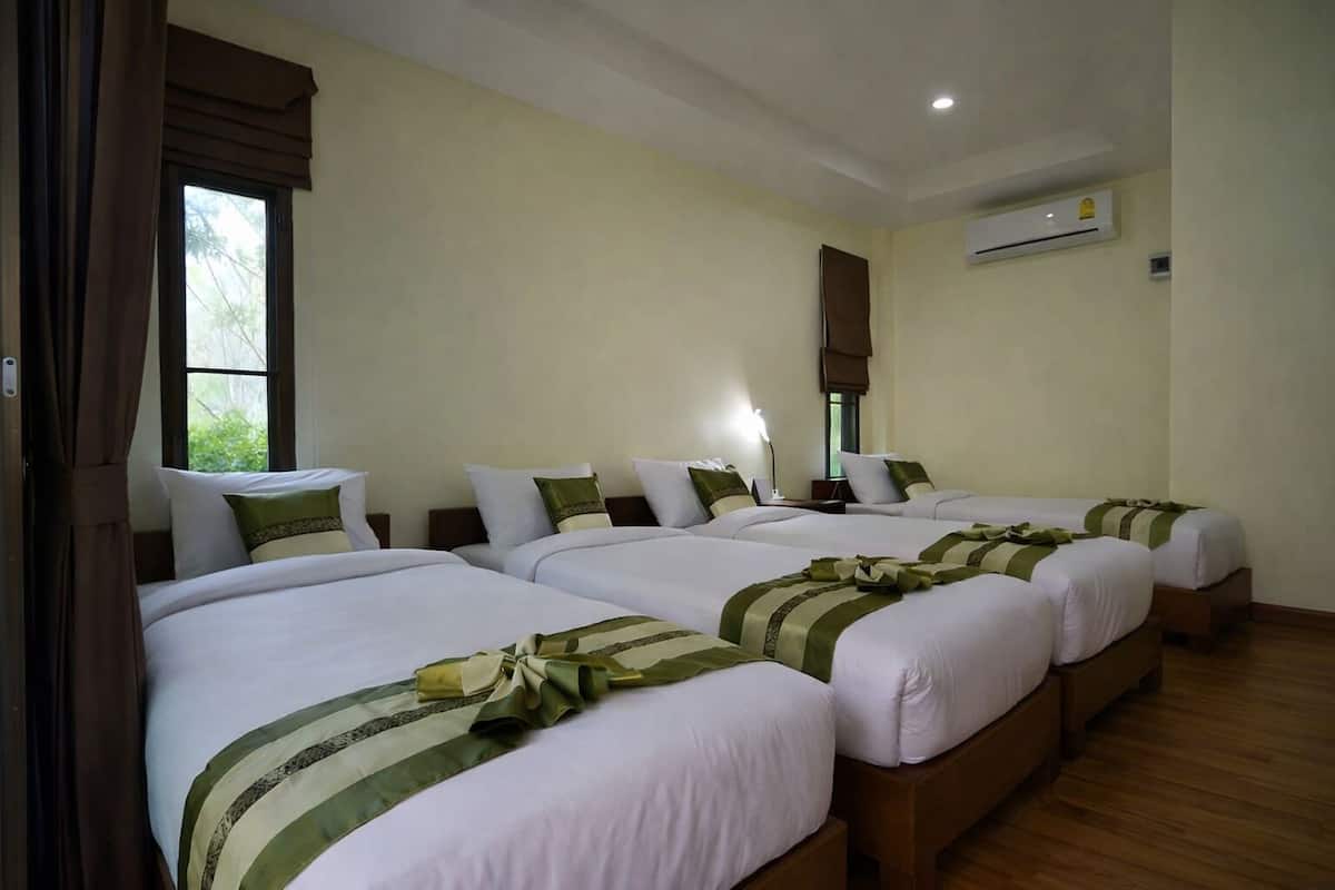 Ein Hotelzimmer mit vier nebeneinander stehenden Betten, jeweils mit weißer Bettwäsche und grünen Akzenten. Es gibt ein Fenster mit Vorhängen, ein Klimagerät an der Wand und einen Holzboden.