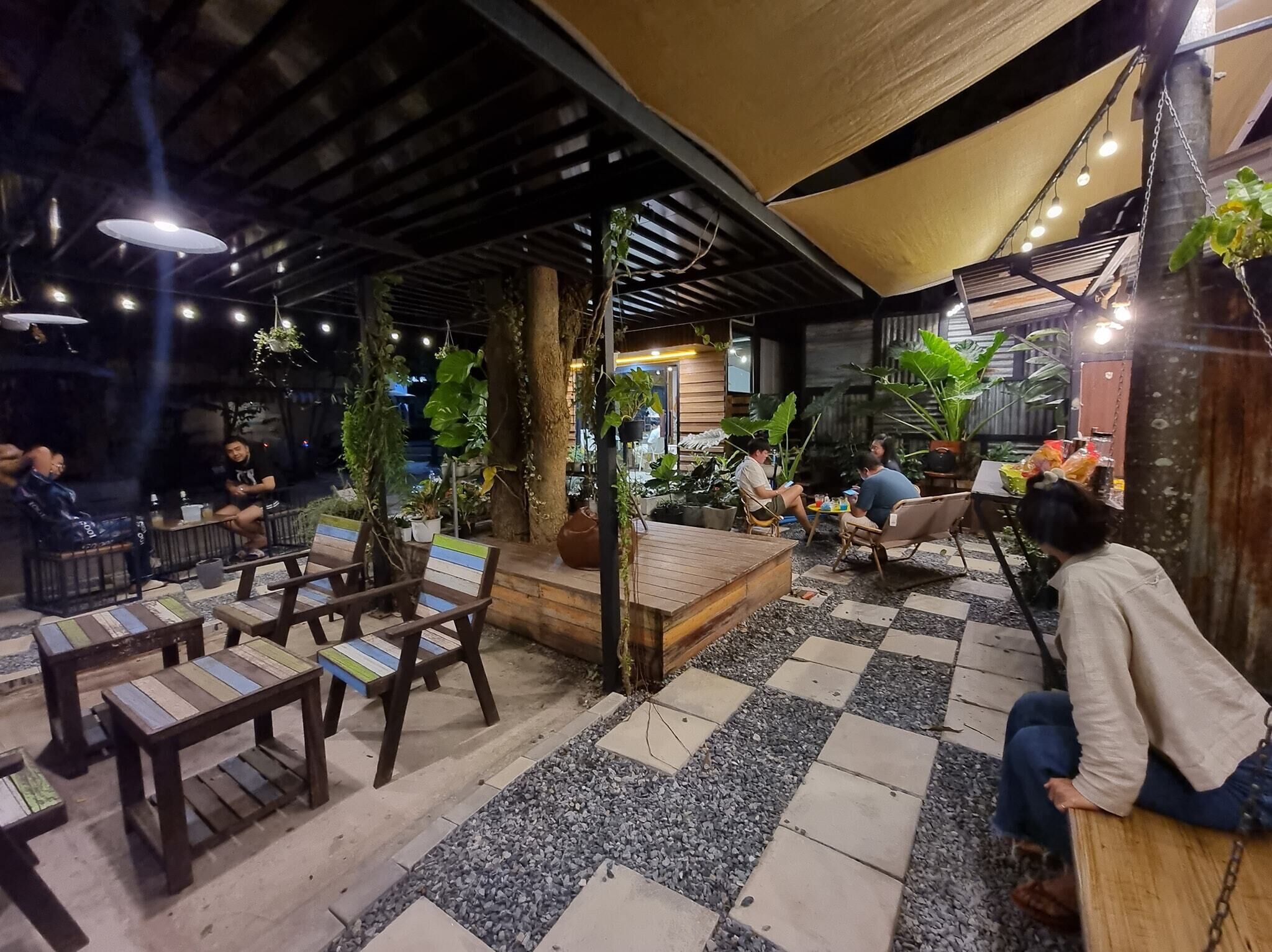 An outdoor seating area with wooden tables and chairs, surrounded by potted plants and a checkered floor pattern.