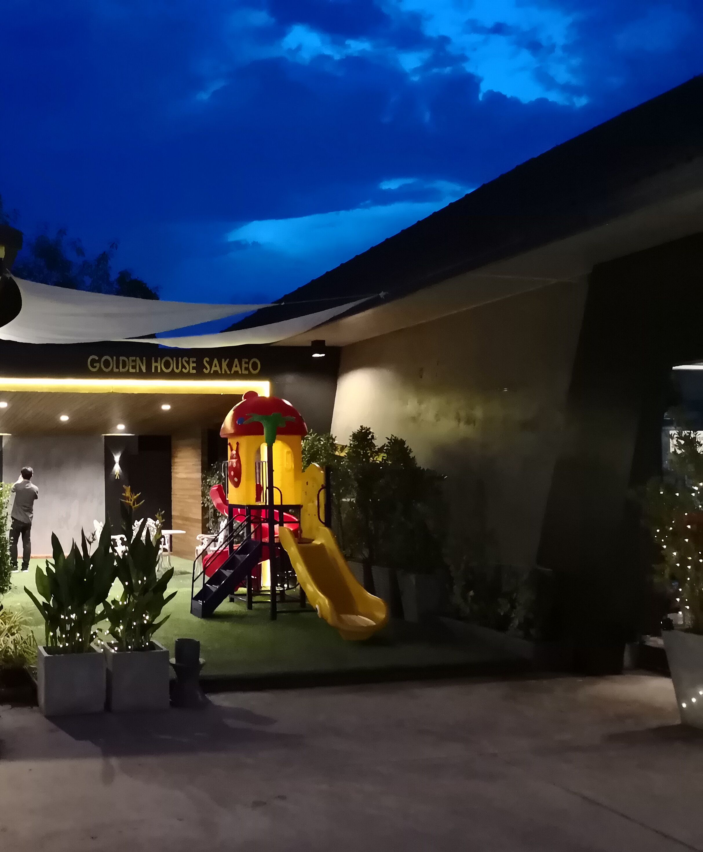 A children's play area with slides and climbing structures, a building entrance labeled "Golden House Sakao," and potted plants around the area.
