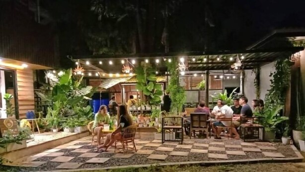 Een terras buiten met tafels en stoelen, omringd door potplanten en lichtslingers.