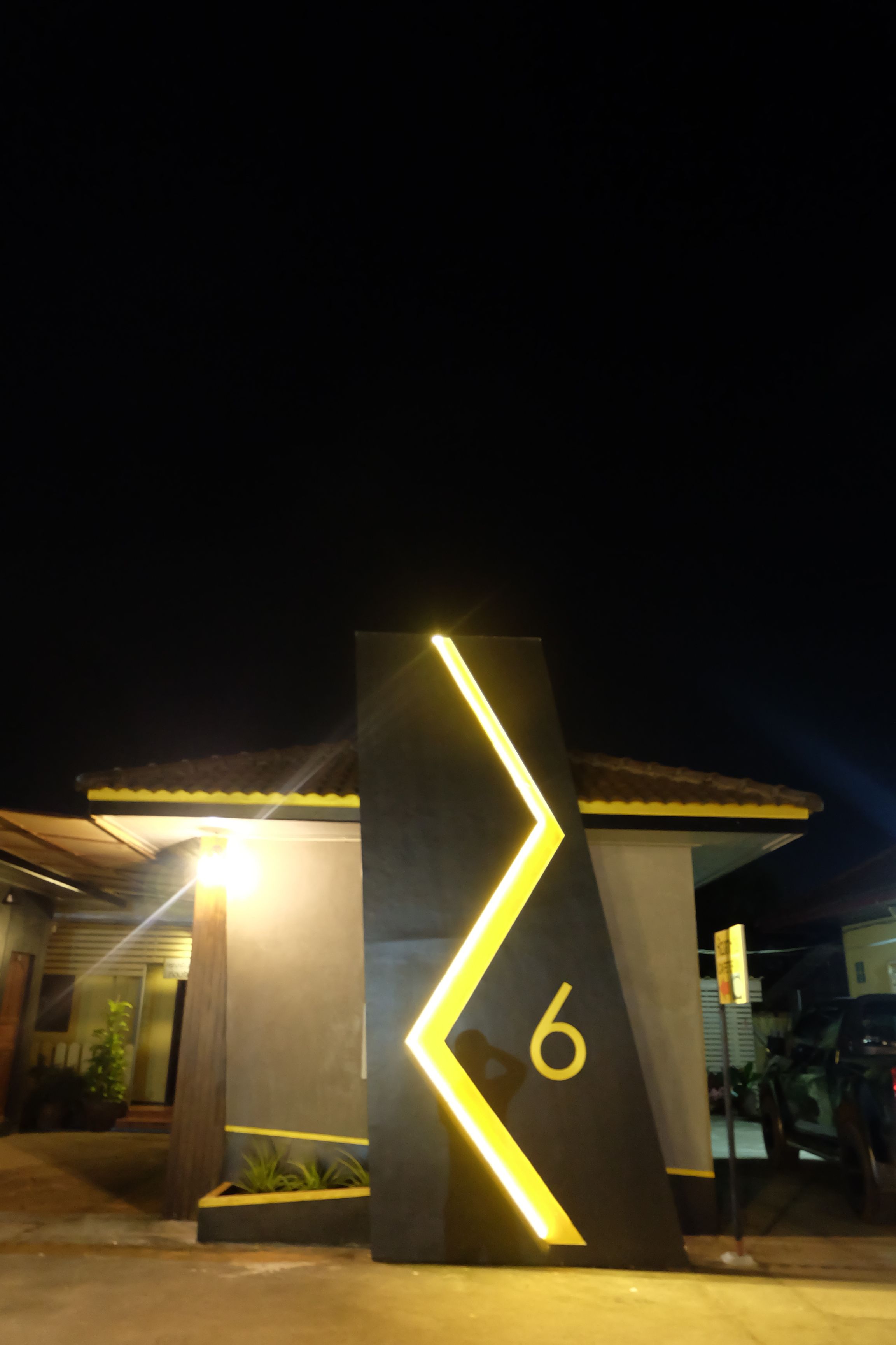 A nighttime view of a building with a prominent number '6' sign, a well-lit entrance, and a parking area visible in the background.