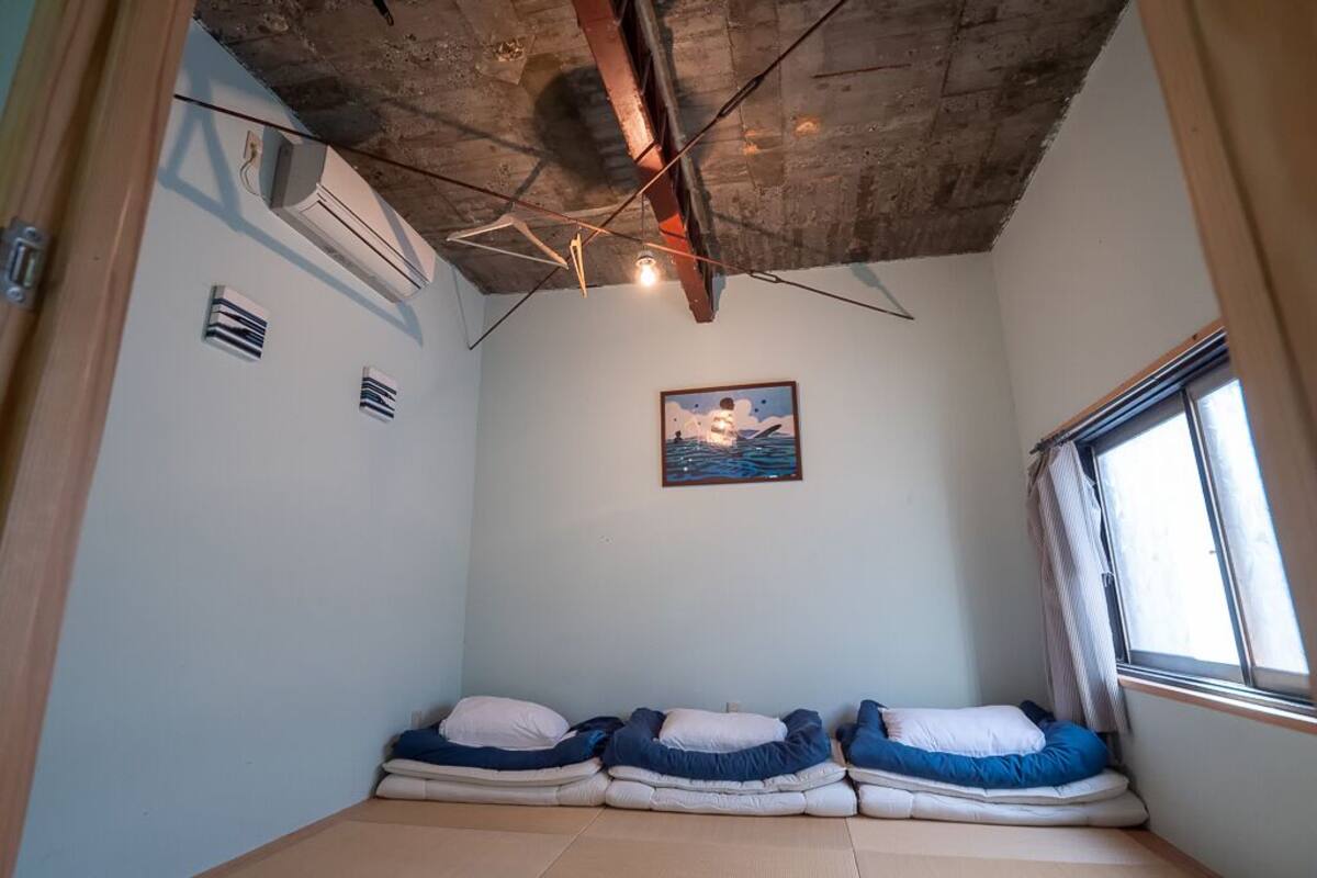 A room with a low ceiling, exposed beams, and a single window. There are two beds with pillows and a painting on the wall.