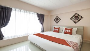 Une chambre d’hôtel avec un grand lit, deux oreillers ornés du mot « OYO », un plaid rouge, une fenêtre avec des rideaux et deux éléments de décoration murale.