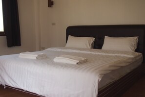 A neatly made bed with white linens and folded towels, a dark headboard, and a plain wall with a window and a light switch.