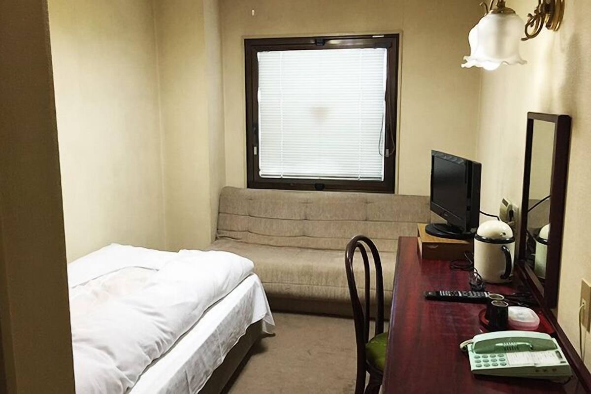 A small hotel room with a bed, a desk with a computer, a telephone, and a window with blinds.