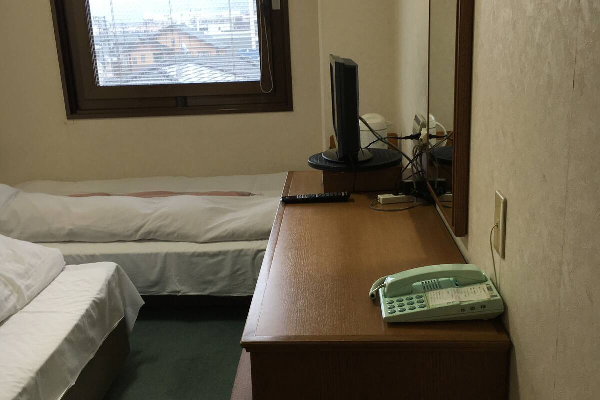 A hotel room with two single beds, a wooden desk with a telephone and a computer monitor, a window with a view of buildings, and a wall-mounted light fixture.