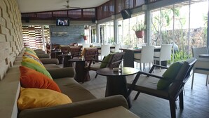 A spacious indoor seating area with a long sofa, multiple tables, and chairs, featuring a television screen and potted plants. - K2 Eco Boutique Lombok (Mataram)