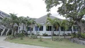 Un bâtiment moderne à l’extérieur blanc et au toit gris, entouré d’un jardin bien entretenu où l’on trouve des palmiers et d’autres plantes tropicales.