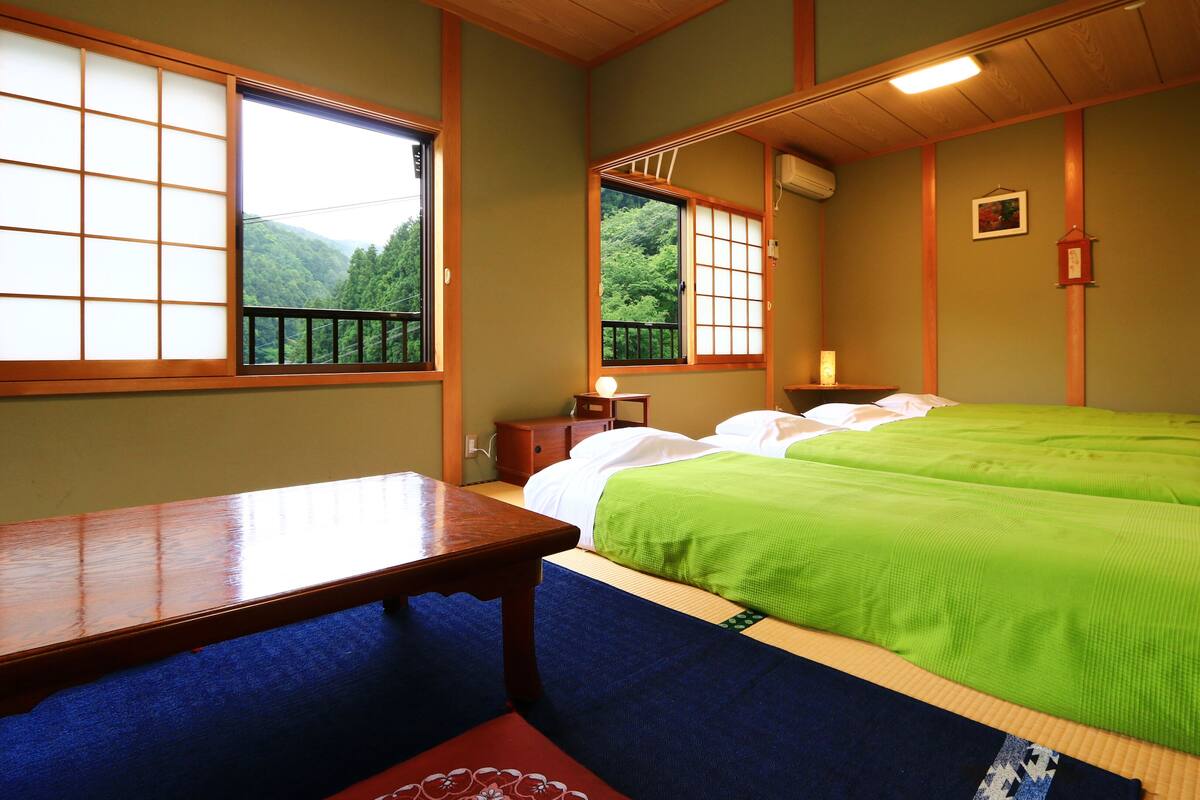 Una habitación tradicional japonesa con suelo de tatami, un escritorio de madera, una cama con ropa de cama verde y vistas a un follaje exuberante a través de ventanas amplias.
