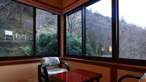 Une pièce d’angle avec une table en bois et deux chaises, de grandes fenêtres donnant sur des arbres et une voie ferrée au loin.