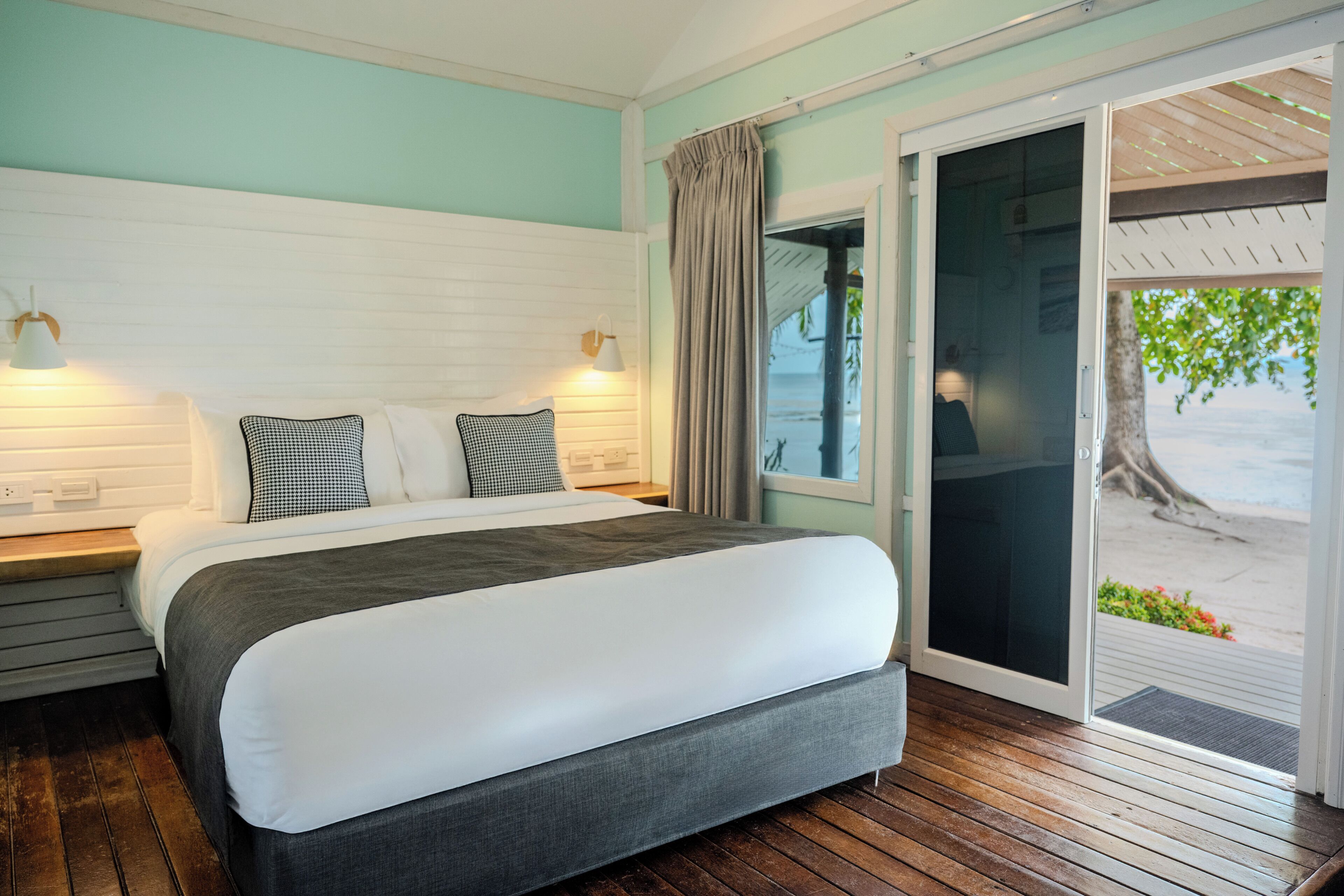 Habitación de hotel con cama, mesita de noche, lámpara y vista a la playa a través de un ventanal.
