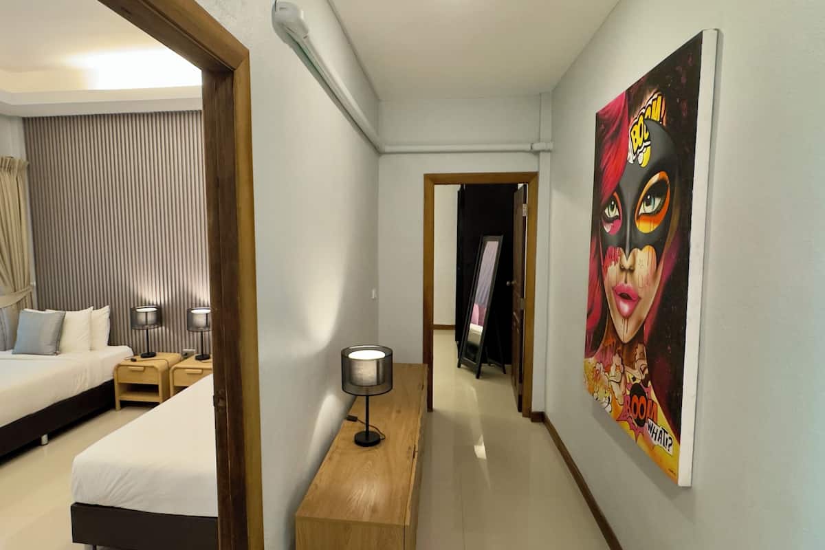 Habitación de hotel con una cama, mesitas de noche, una lámpara, un cuadro y un pasillo que conduce a otra habitación.
