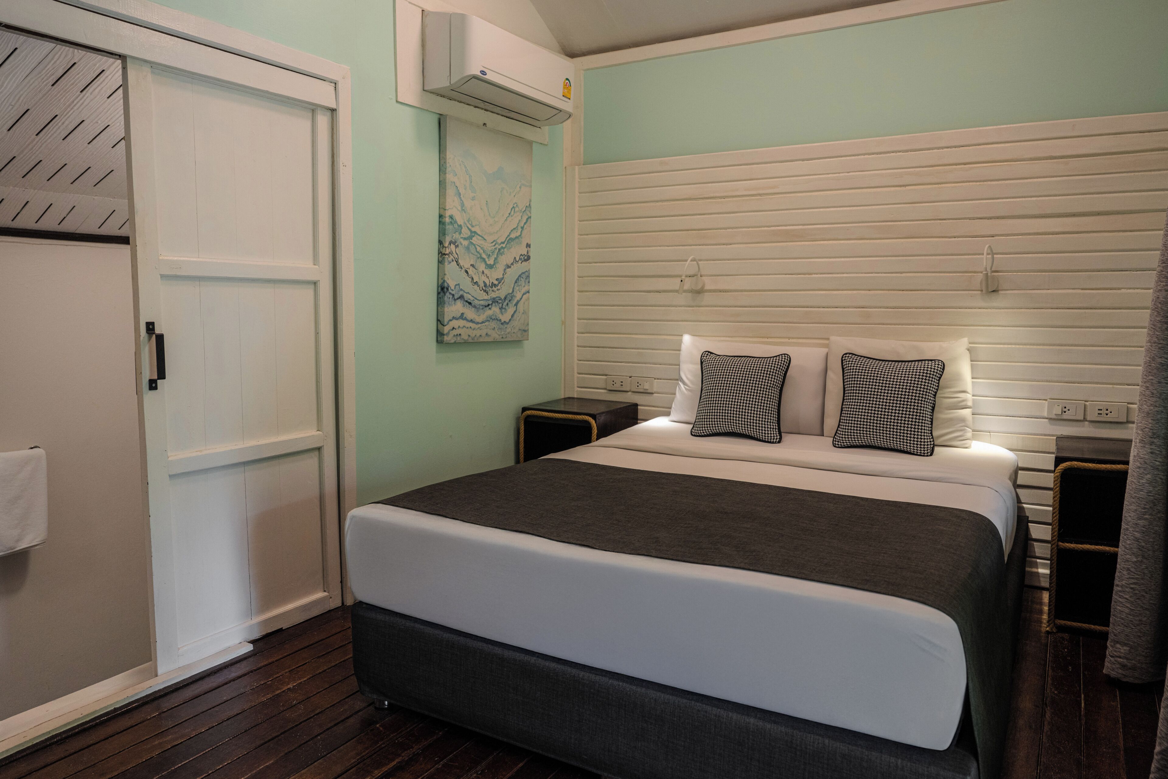 Une chambre à coucher comprenant un lit, une table de chevet, un climatiseur fixé au mur, une porte et un tableau.