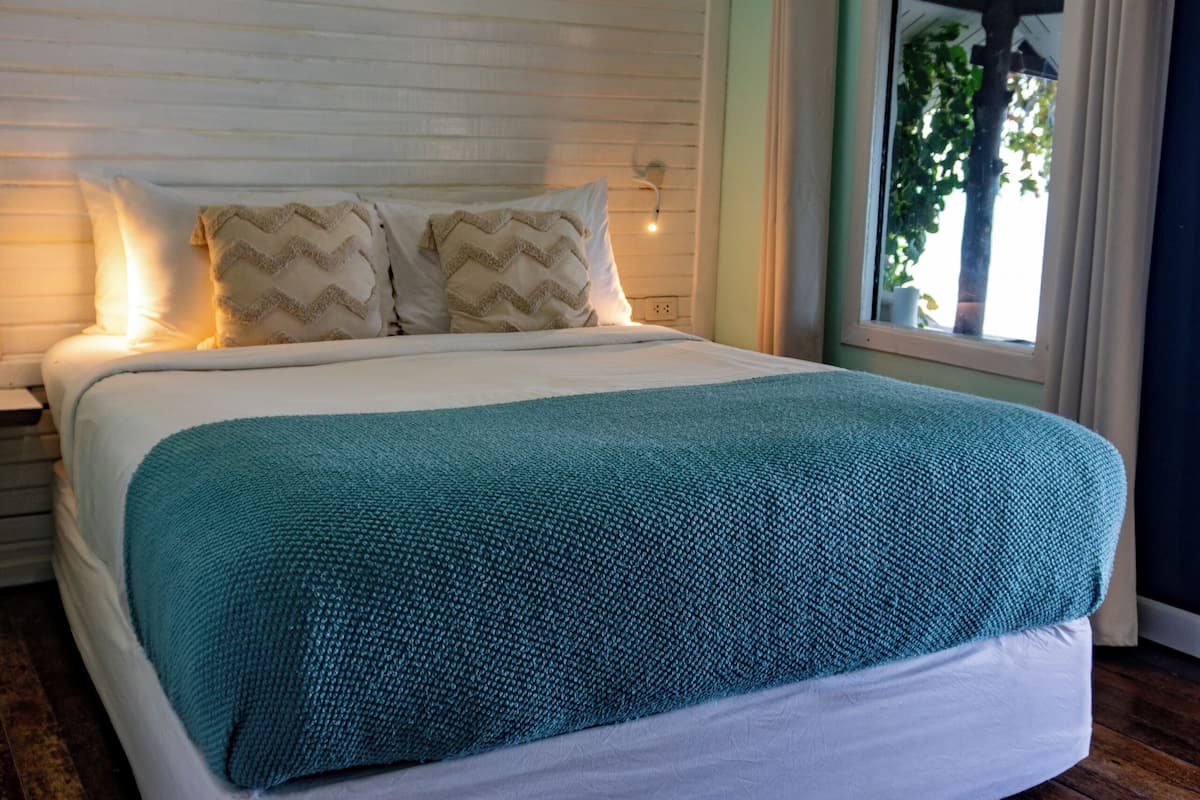 Un dormitorio con cama, almohadas, ventana, cortinas y suelo de madera.