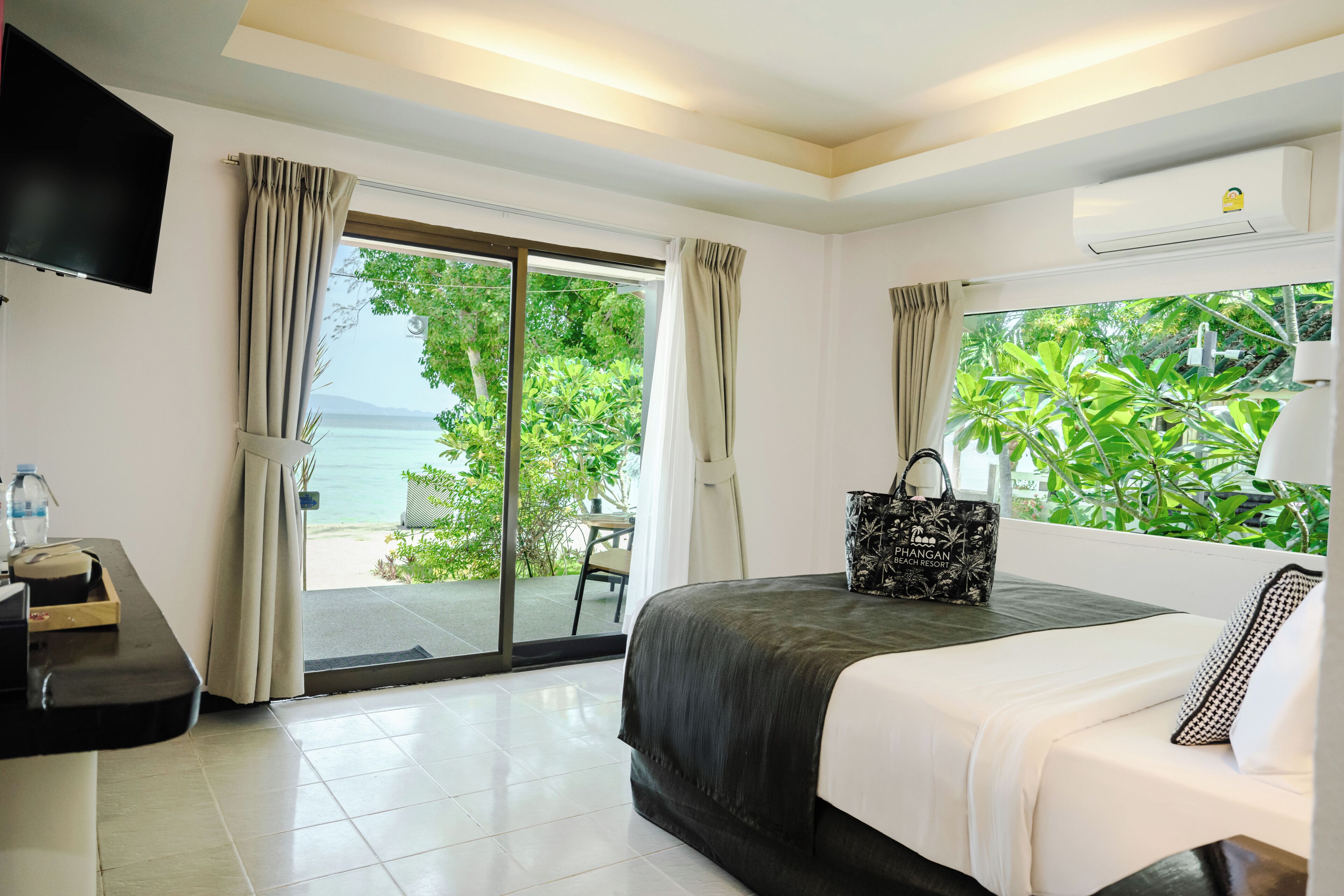 A hotel room with a bed, a desk, a TV, a window with a view of the sea, and an air conditioning unit.
