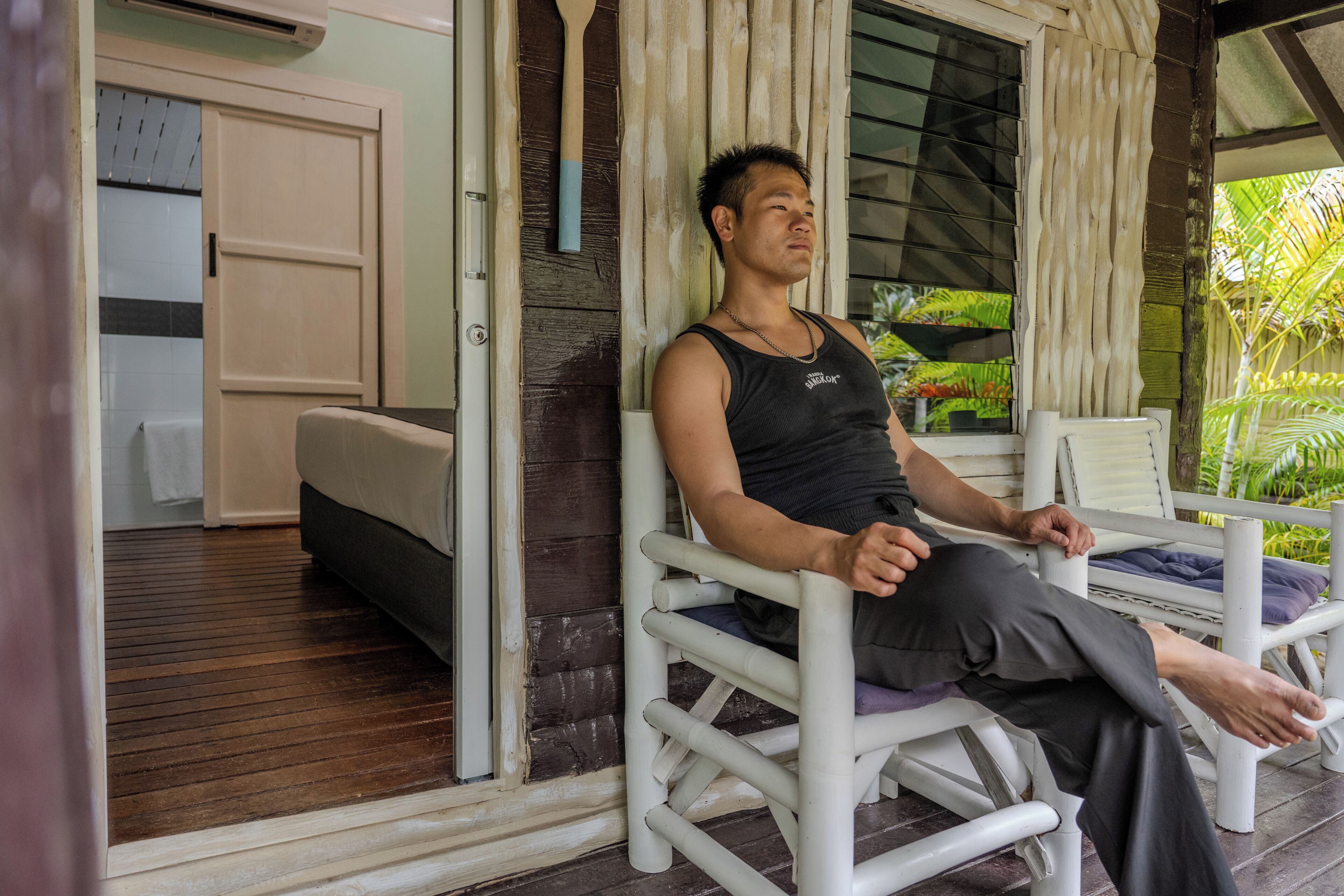 Une personne est assise sur une chaise blanche dans une pièce dont le sol est en bois, qui contient un lit et une fenêtre avec des rideaux.