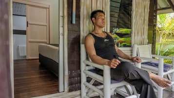Une personne est assise sur une chaise blanche dans une pièce dont le sol est en bois, qui contient un lit et une fenêtre avec des rideaux.