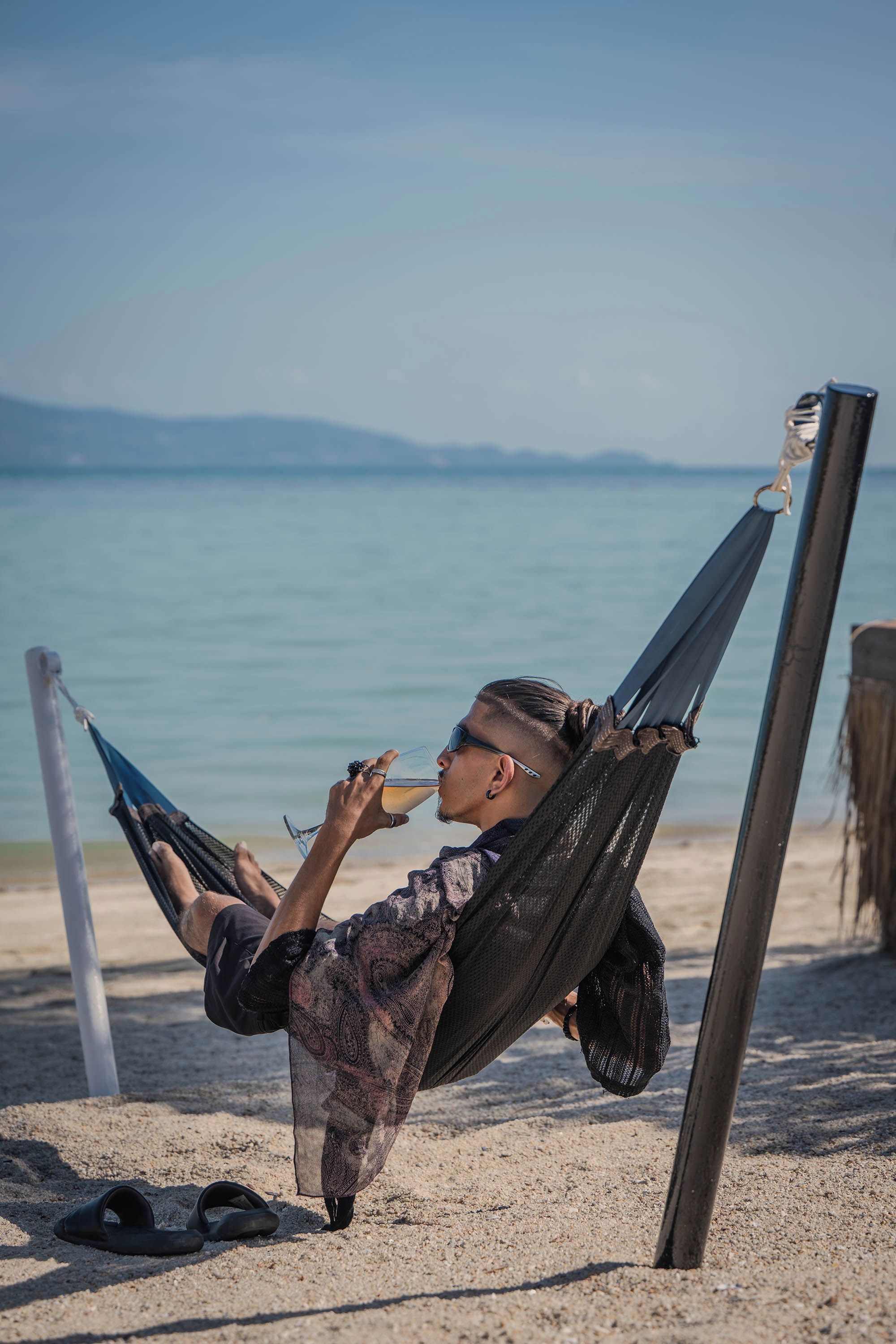 A hammock on a sandy beach with a clear view of the sea and distant mountains.