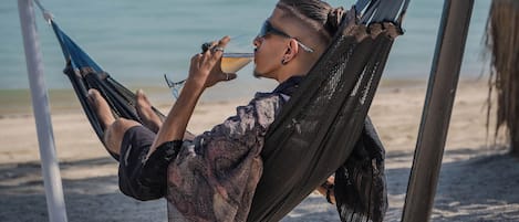 A hammock on a sandy beach with a clear view of the sea and distant mountains.