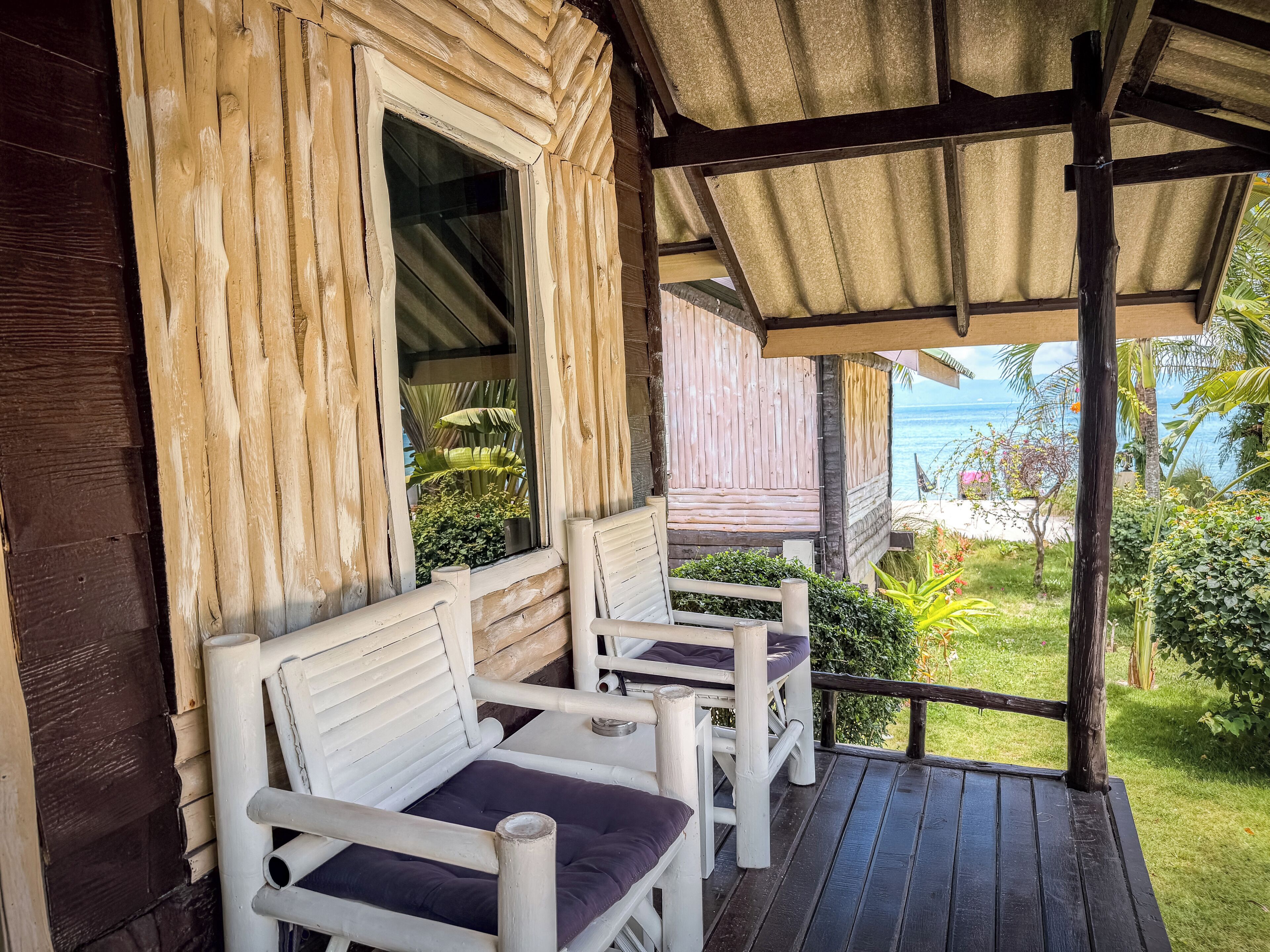 Een houten veranda met twee witte stoelen en een bankje, met uitzicht op een weelderige tuin en de zee.