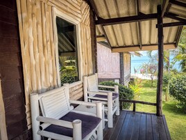 A wooden porch with two white chairs and a bench, overlooking a lush garden and the sea.
