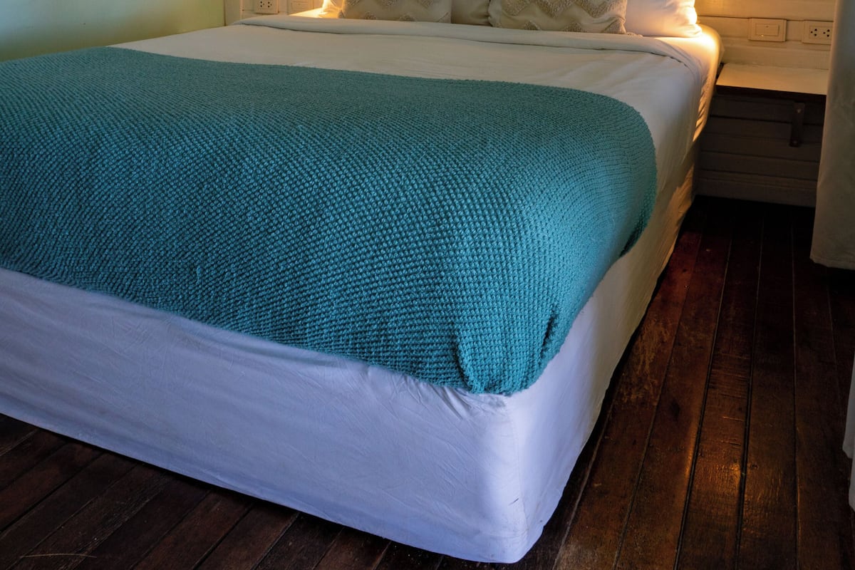 Una cama con cabecera blanca y ropa de cama color turquesa, un aire acondicionado montado en la pared y piso de madera.