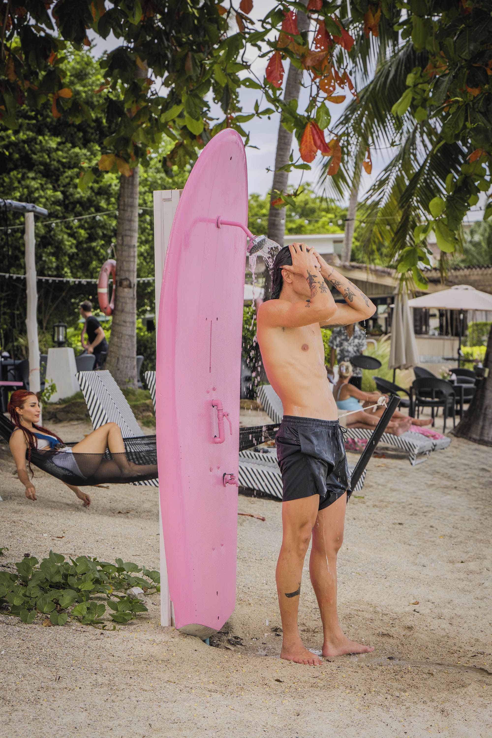 A person standing next to a pink surfboard on a sandy beach with palm trees and a hammock in the background.