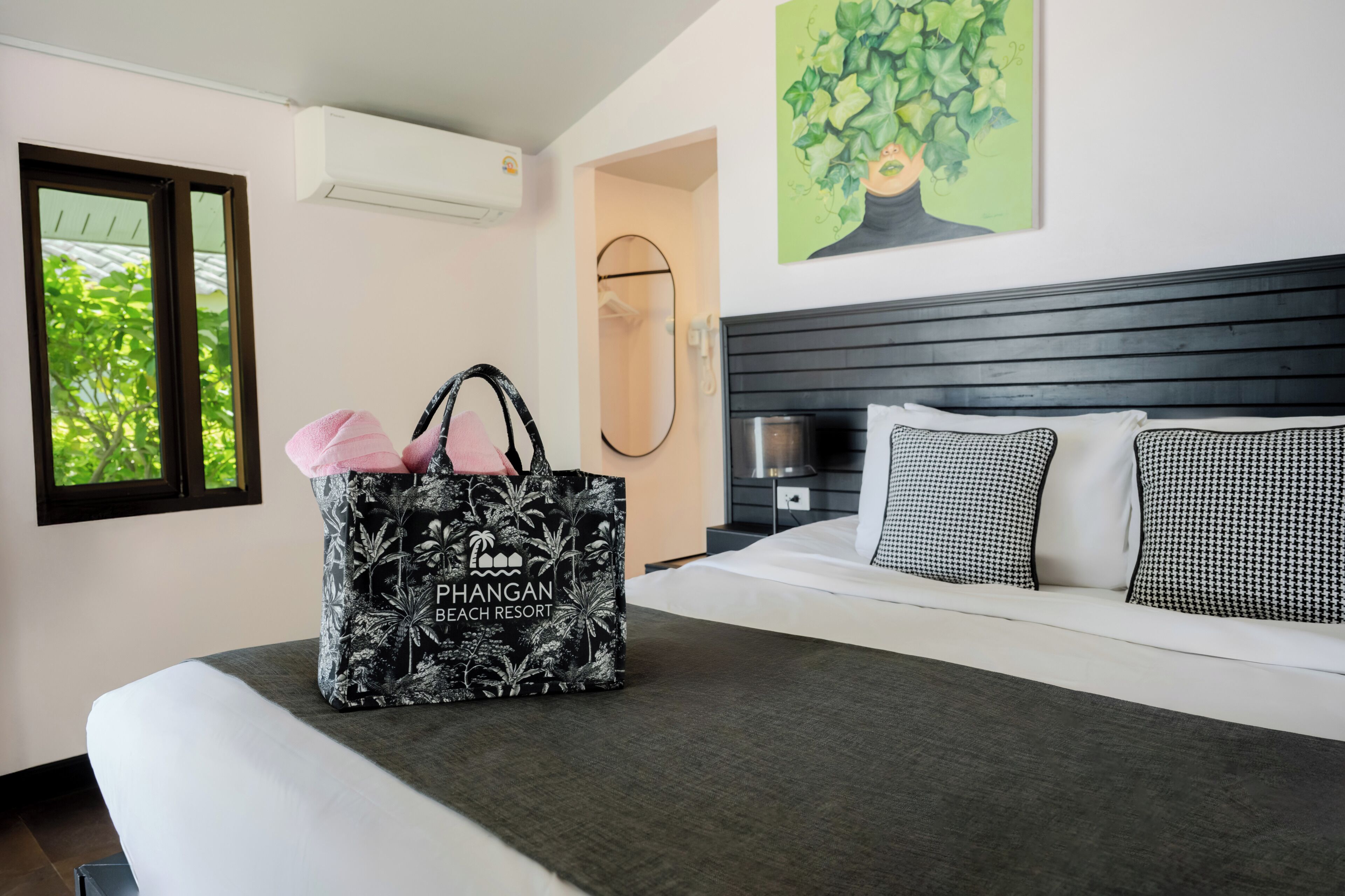 Ein Hotelzimmer mit einem Bett, einer großen, schwarz-weiß gemusterten Tasche, einem Fenster mit Blick auf Bäume und einem Baumgemälde an der Wand.