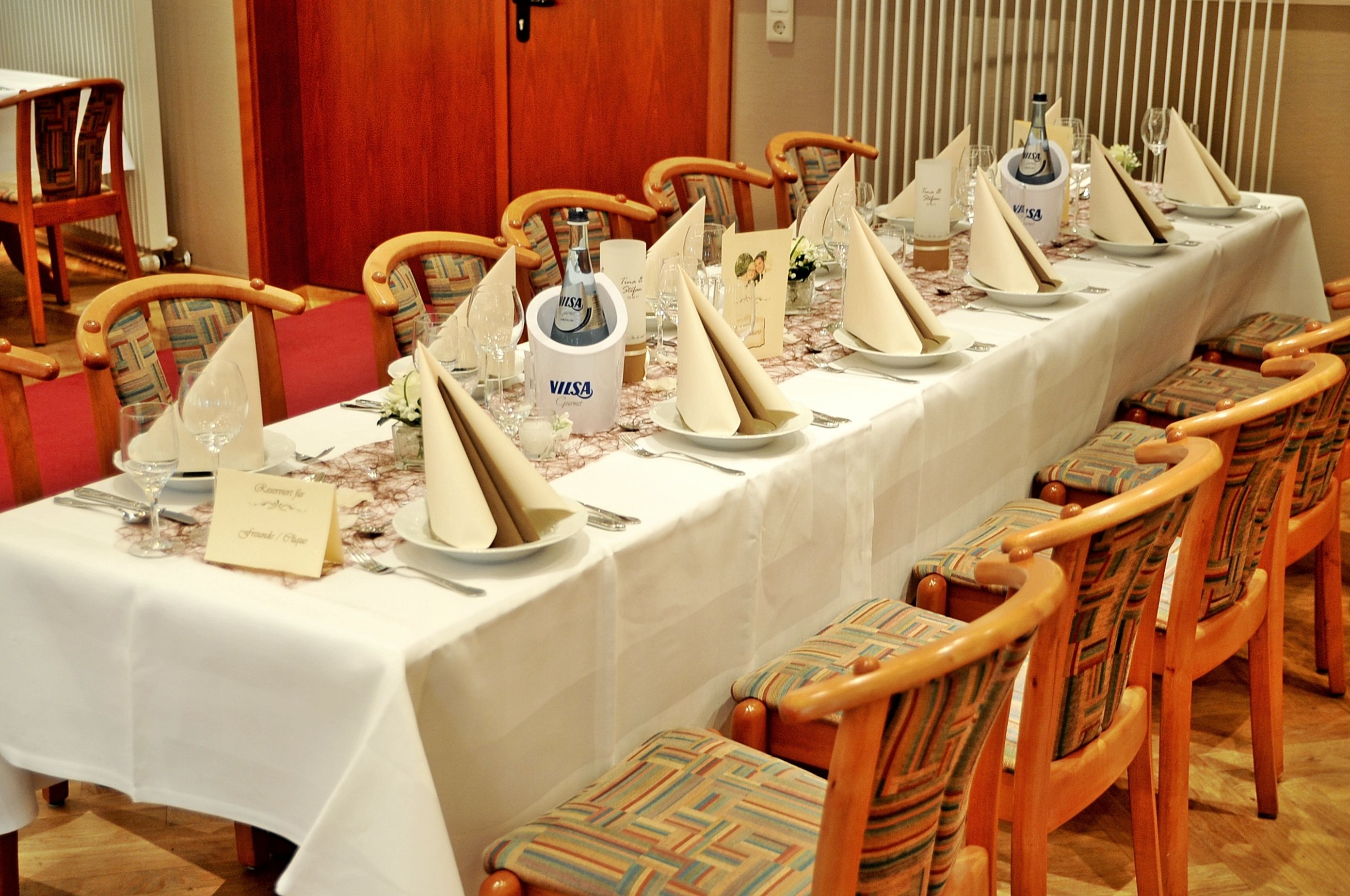 A long dining table set for a formal meal with multiple chairs, a white tablecloth, and V.I.S.A. branded champagne.