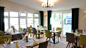 A dining area with multiple tables and chairs, large windows, and a chandelier. - Hotel Restaurant Burgerklause Tapken (Garrel)