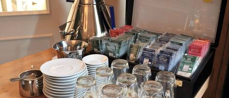 Una mesa con pilas de platos blancos, tazas de vidrio transparente y un hervidor de plata.