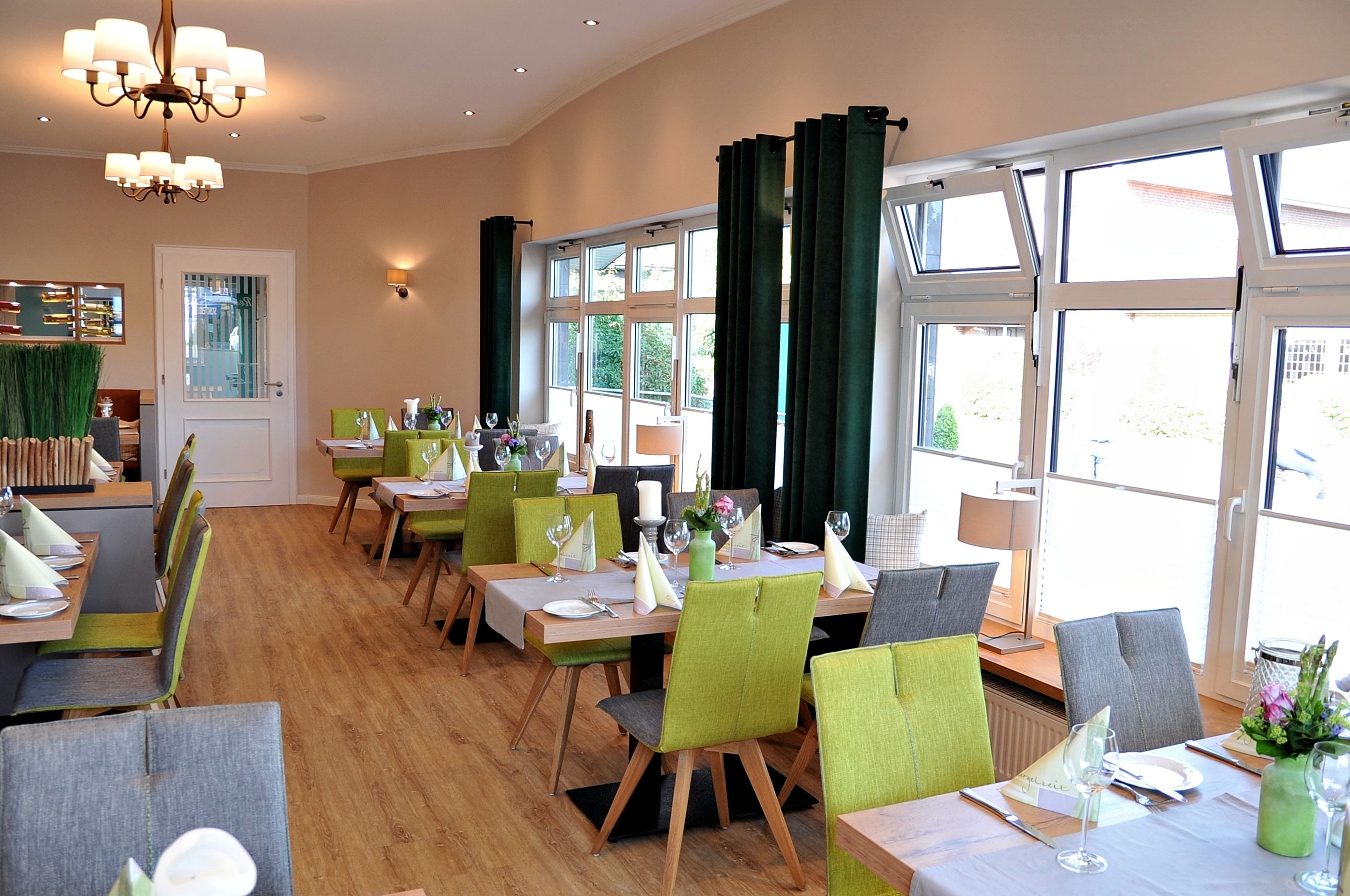 A restaurant with multiple tables, green and grey chairs, and large windows with curtains.