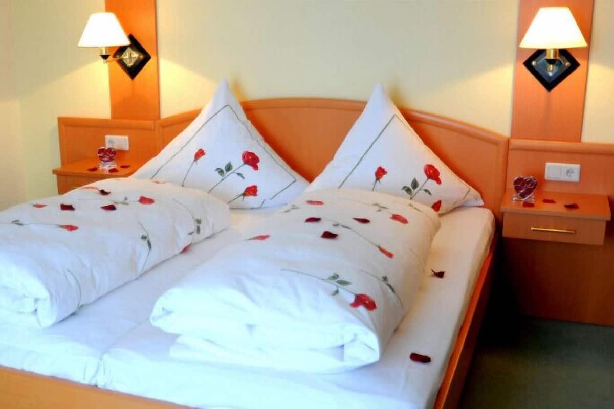 A double bed with white bedding and rose patterns, flanked by two bedside tables with lamps and a small decorative item on each.