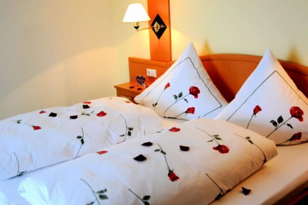 A bed with white bedding featuring red rose patterns, a wooden headboard, and a nightstand with a lamp and a mirror.