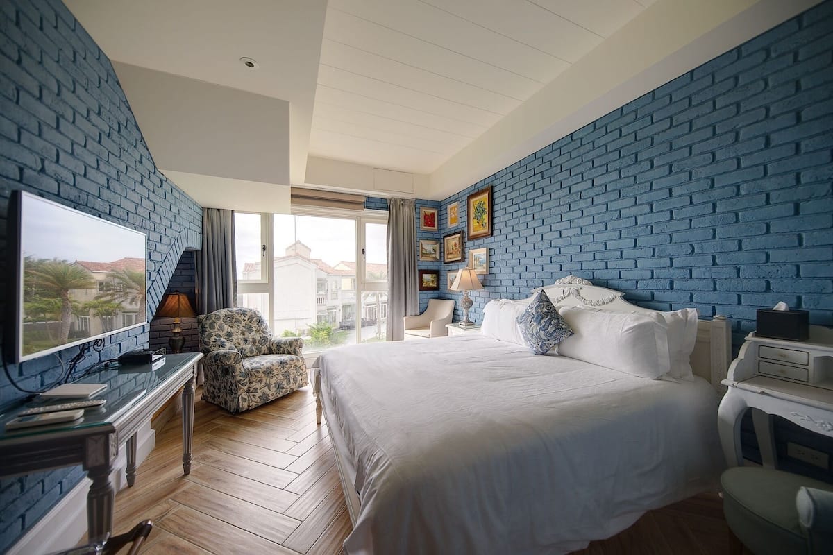 A hotel room with a brick-patterned wall, a large bed, a TV, a desk, and a chair.