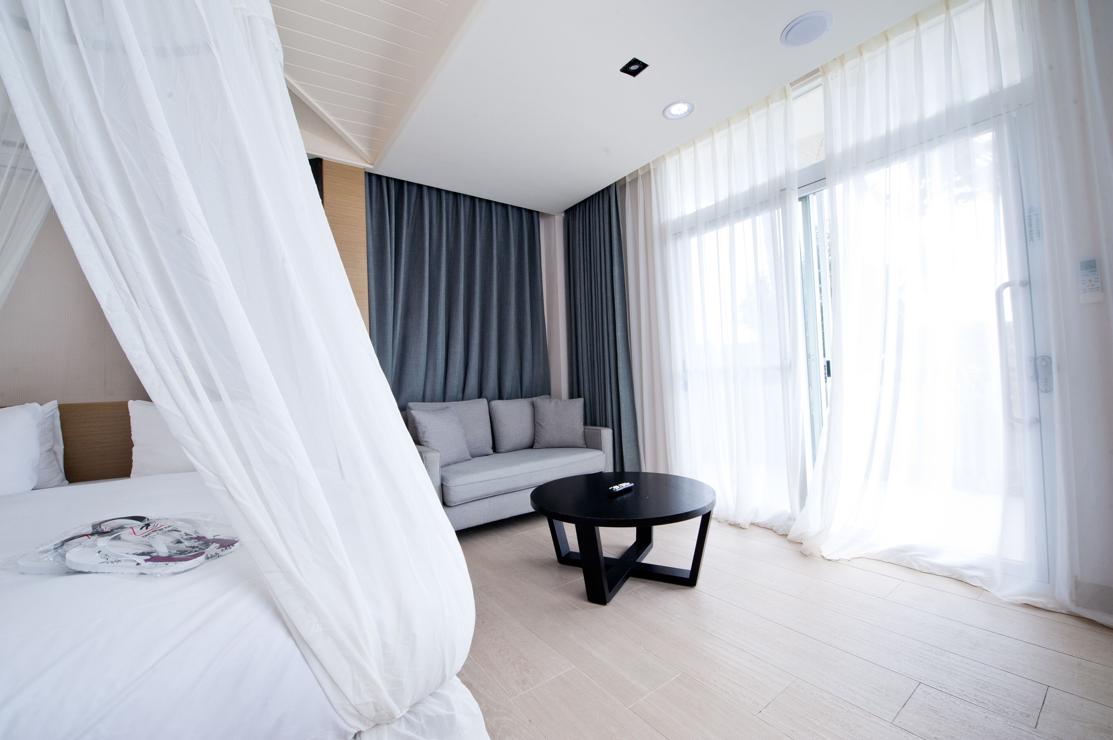 A modern hotel room with a bed, a grey sofa, a round black table, and sheer curtains.