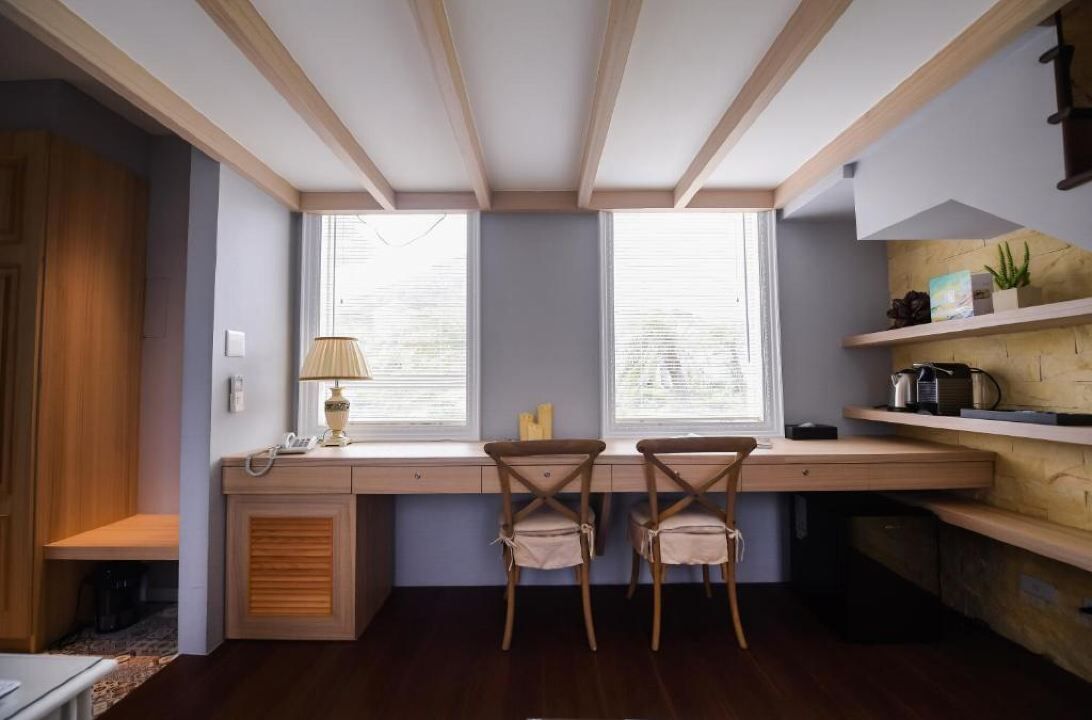 A compact kitchen with a built-in desk and two chairs, a window with blinds, and wooden shelves with kitchen items.