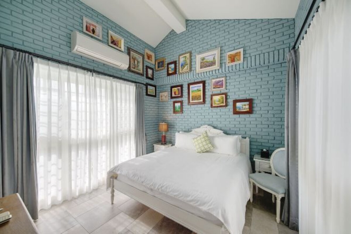 A bedroom with a blue brick wall, a bed, a bedside table, a chair, and a window with curtains.