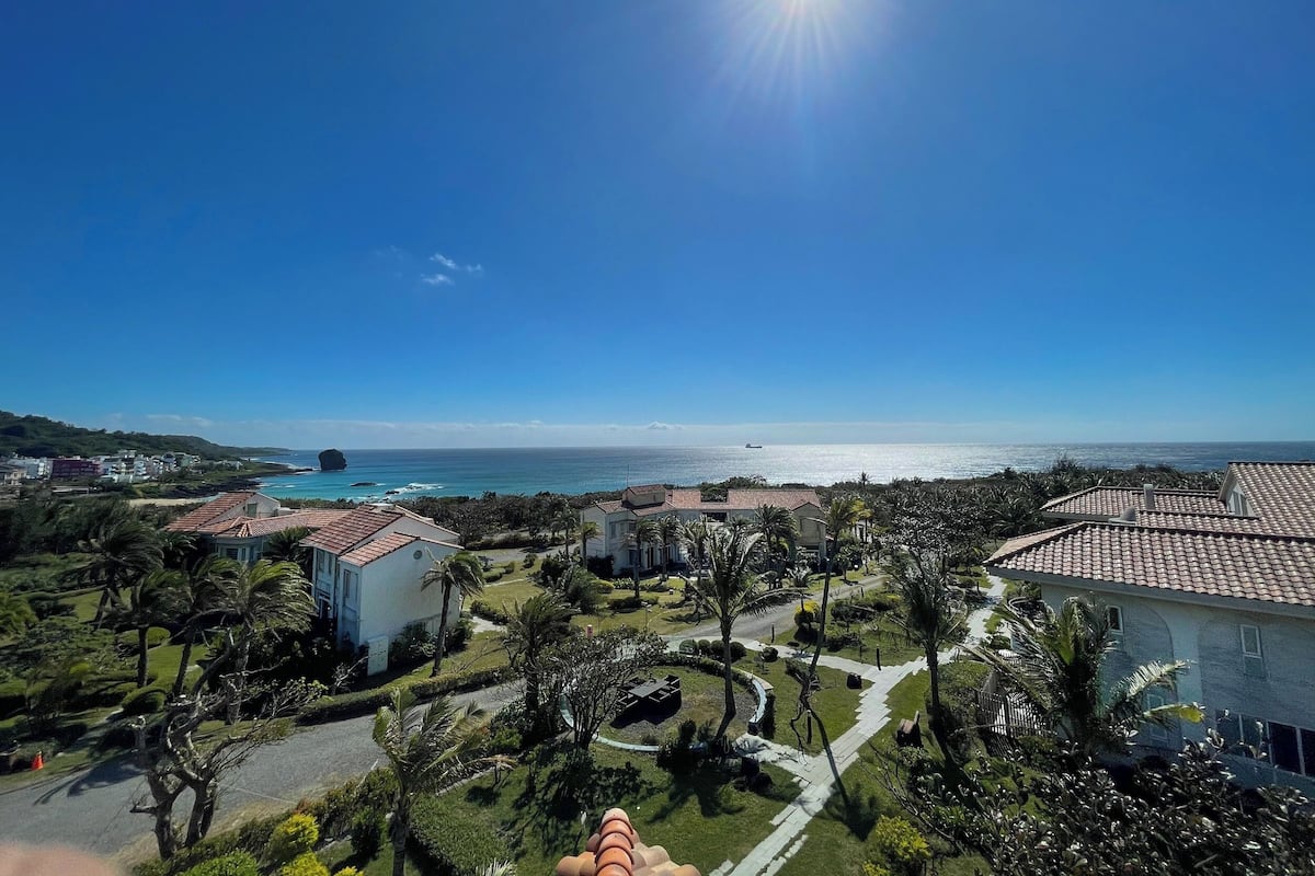 A coastal resort with terracotta-roofed buildings, a clear blue sky, and a view of the ocean.
