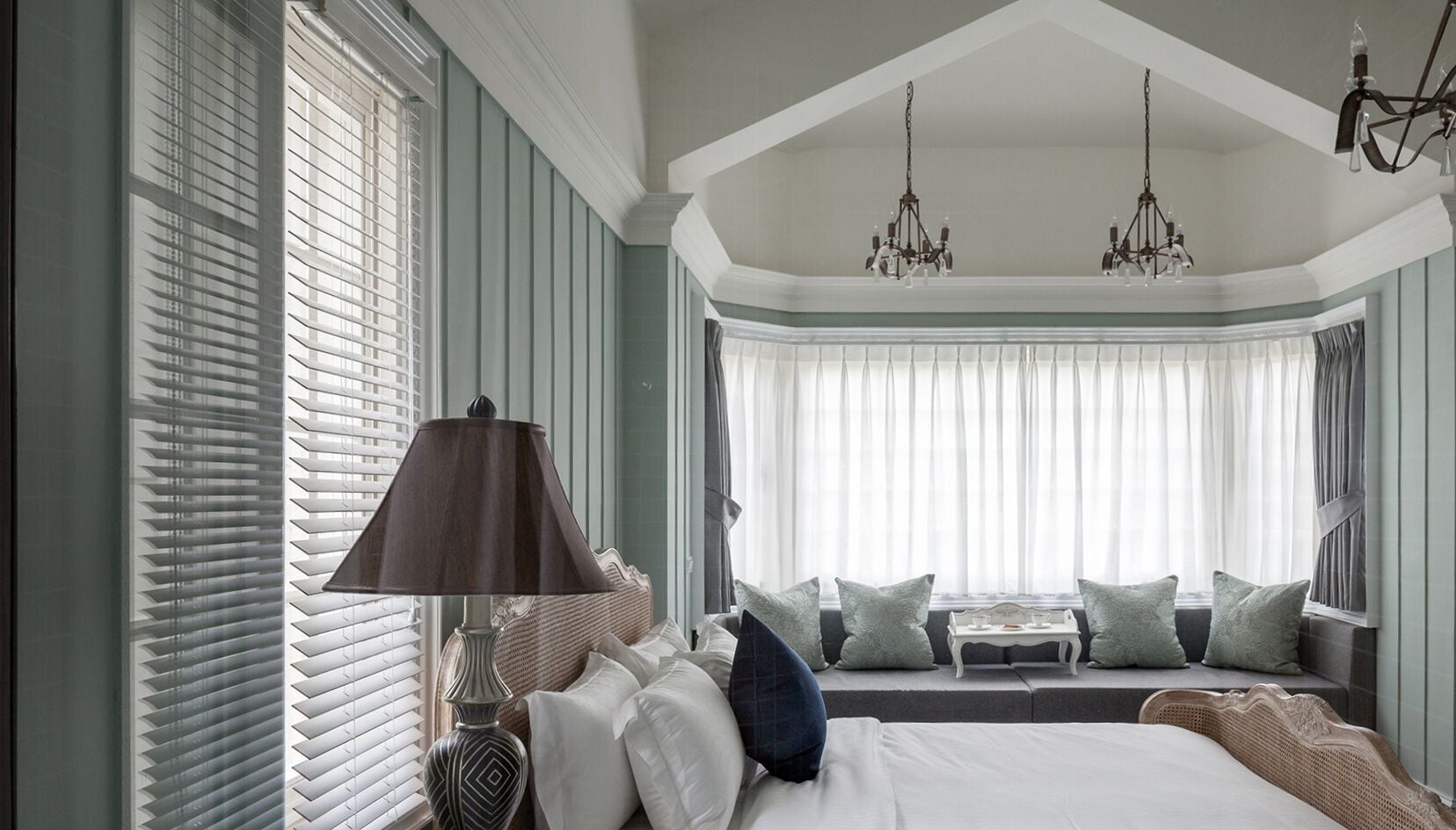 Un dormitorio con una cama grande, una lámpara de noche, un asiento junto a la ventana con cojines y dos candelabros.