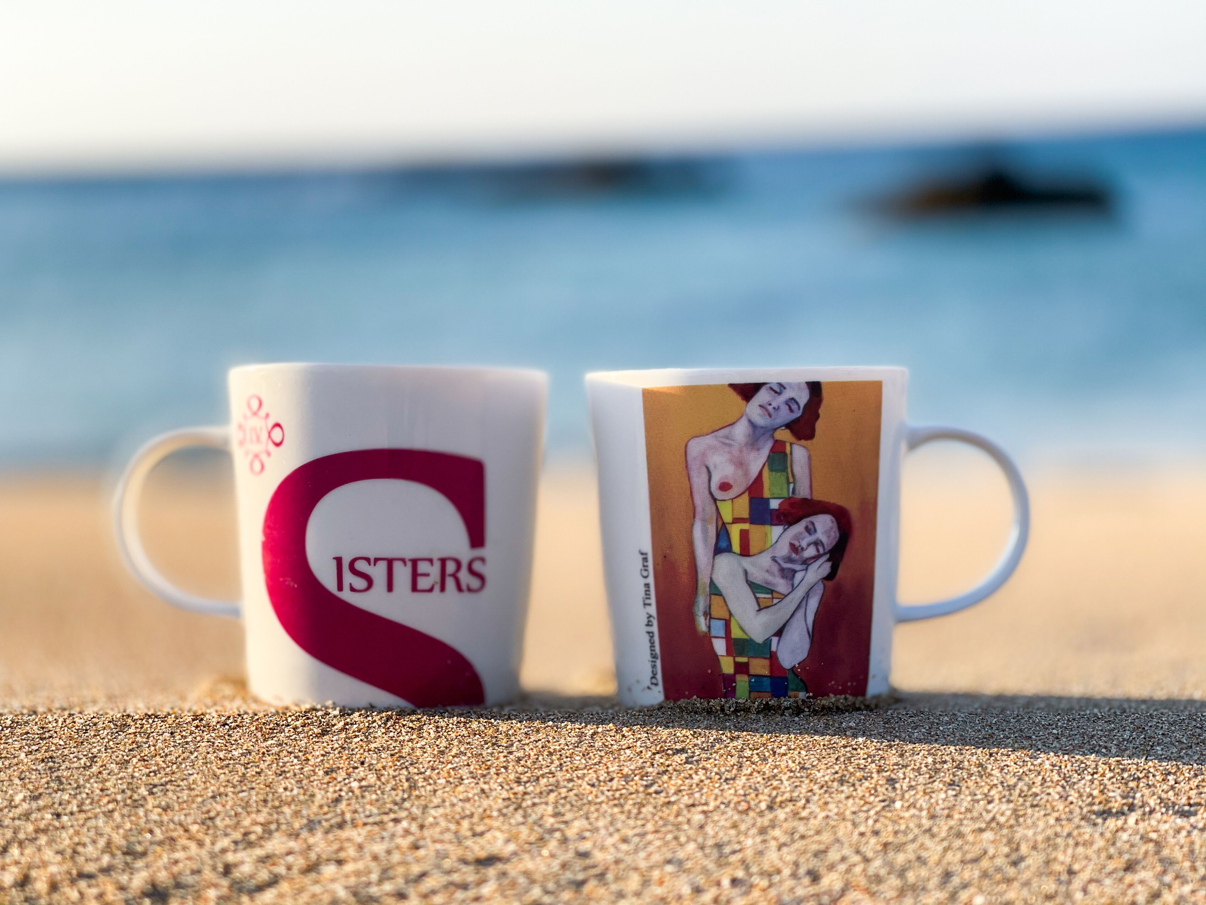 Two coffee cups on a sandy surface with a blurred ocean background.