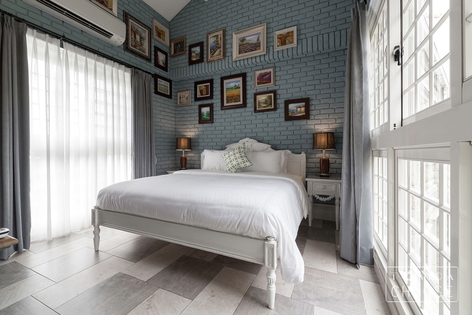 A bedroom with a white bed, grey curtains, and a brick-patterned wall with framed pictures.