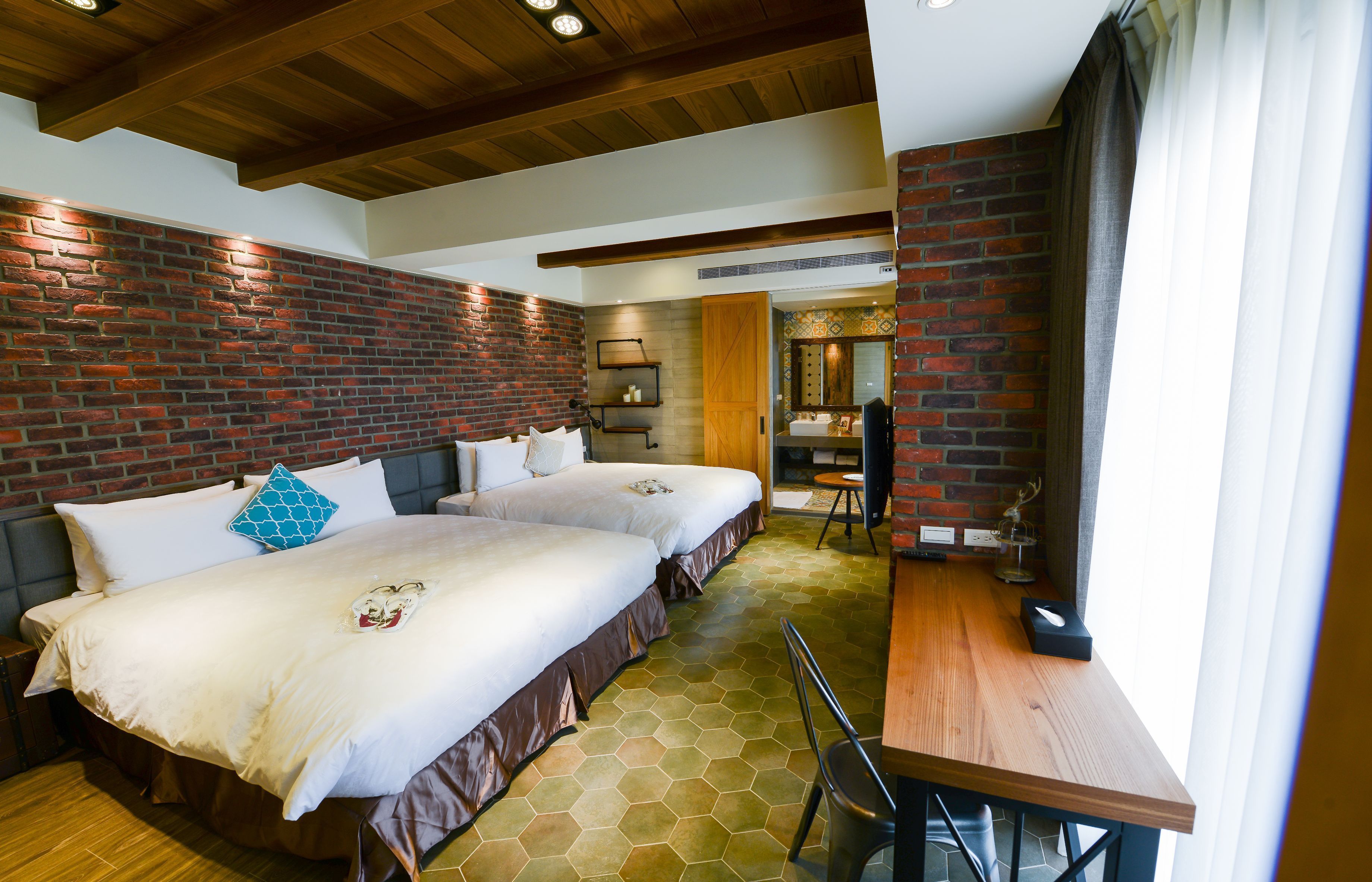 A hotel room with two beds, a wooden table, and a brick wall.