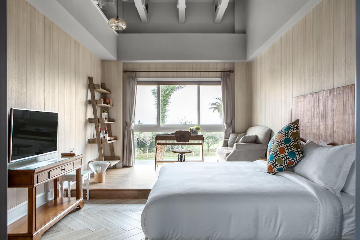 A bedroom with a large bed, a TV on a wooden stand, a desk with a chair, and a window with a view of palm trees.