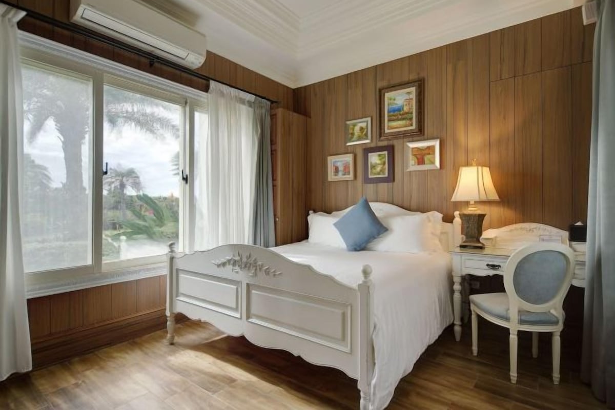 Un dormitorio con cama, escritorio y silla. Hay ventanas con cortinas, una lámpara y cuadros enmarcados en la pared.